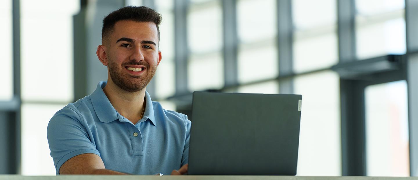 A young man with a laptop smiling at the camera